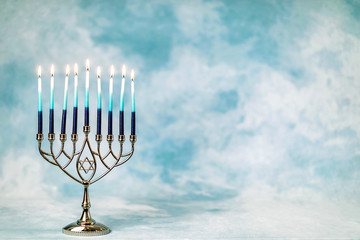 A silver menorah for the Jewish holiday Hanukkah with burning glowing eight candles on fire lit up on a blue background with copy space