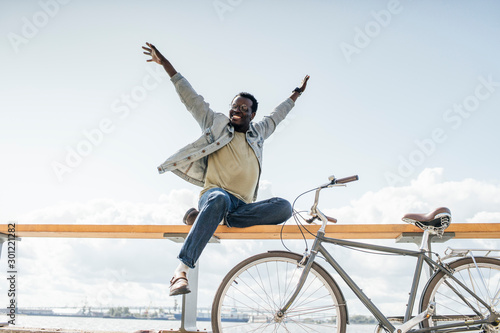 Young man with bicycle, sitting on railing by the sea, pretending to fly photo