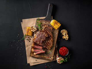 Fried steaks with herbs and spices on wooden board, parchment, grilled garlic, corn, sauce in dark bowl on black background. Close-up. Top view.