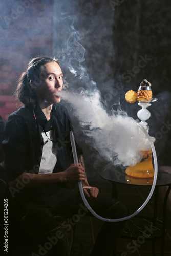 Portrait of man smoking traditional hookah pipe and making smoke clouds with shisha. Man exhaling smoke in hookah cafe or lounge