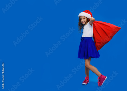 Full-lenght pretty little curly smiling girl wearing in santa hat and skirt carrying red bag, over on blue background.
