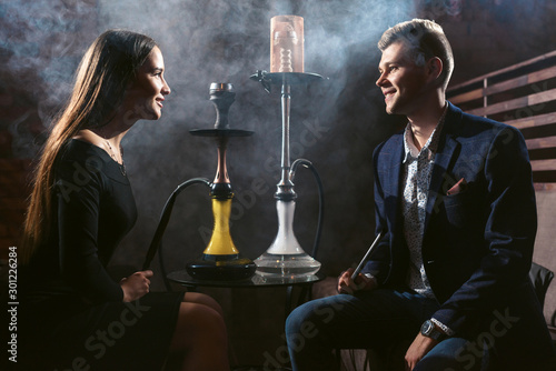 Beautiful couple woman and man in backlight in love smoking hookah and drinks cocktails in the bar