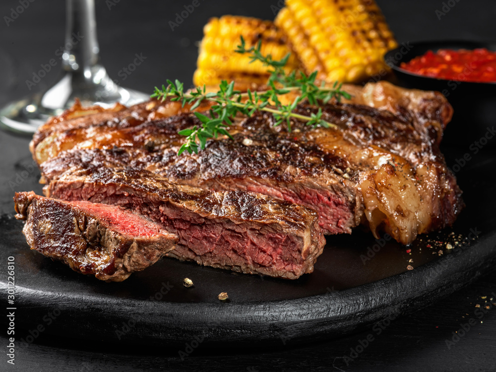 Sliced, fried, spiced juicy steaks with herbs on a dark board, grilled corn, red sauce and glass on a black background. Close-up shot. Side view.