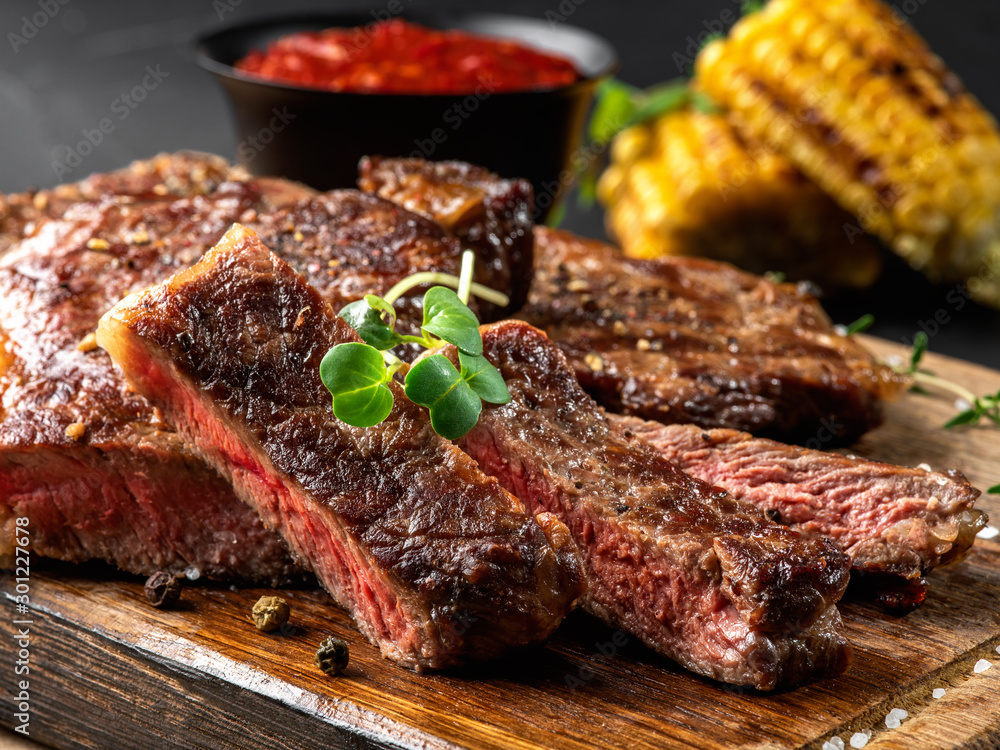 Sliced, fried, spiced steaks with herbs on wooden board, grilled corn, red sauce in small dark bowl on a black background. Close-up shot. Side view.