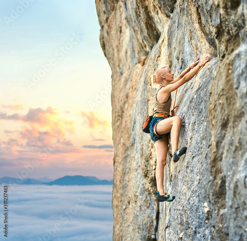 side view of young athletic woman rock climber climbing on the cliff. a woman climbs on a vertical rock wall with sunset sky on background. Conquering, overcoming and active lifestyle concept.