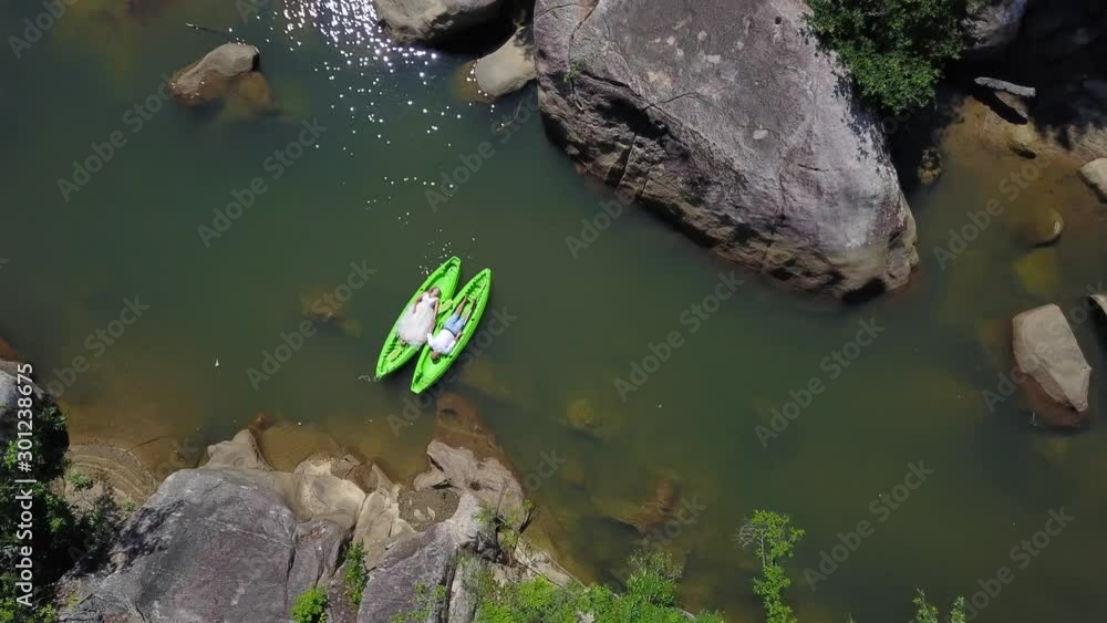 A flying camera shoots from top to bottom as the bride and groom swim in two small boats among the jungle and large stones.