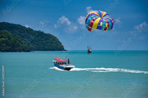 Parasailing on the waves of the azure Andaman sea under the blue sky near the shores of the sandy beautiful exotic and stunning Cenang beach in Langkawi island, photo