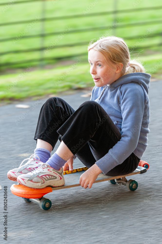 Excited young blonde girl sitting on a fast moving skateboard. Panning action shot with motion blurred background Excited young blonde girl sitting on a fast moving skateboard. Panning action shot with motion blurred background