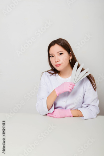 Portrait of a girl, master of nails in a white coat and gloves sitting at a table on a white background with nail files in her hands.