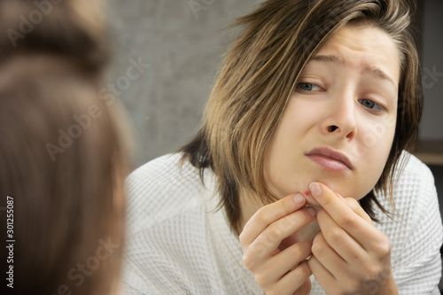 A woman in the bathroom  looking at herself in the mirror and trying to squeeze a pimple  zit with a sad  scared  disappointed facial expression.