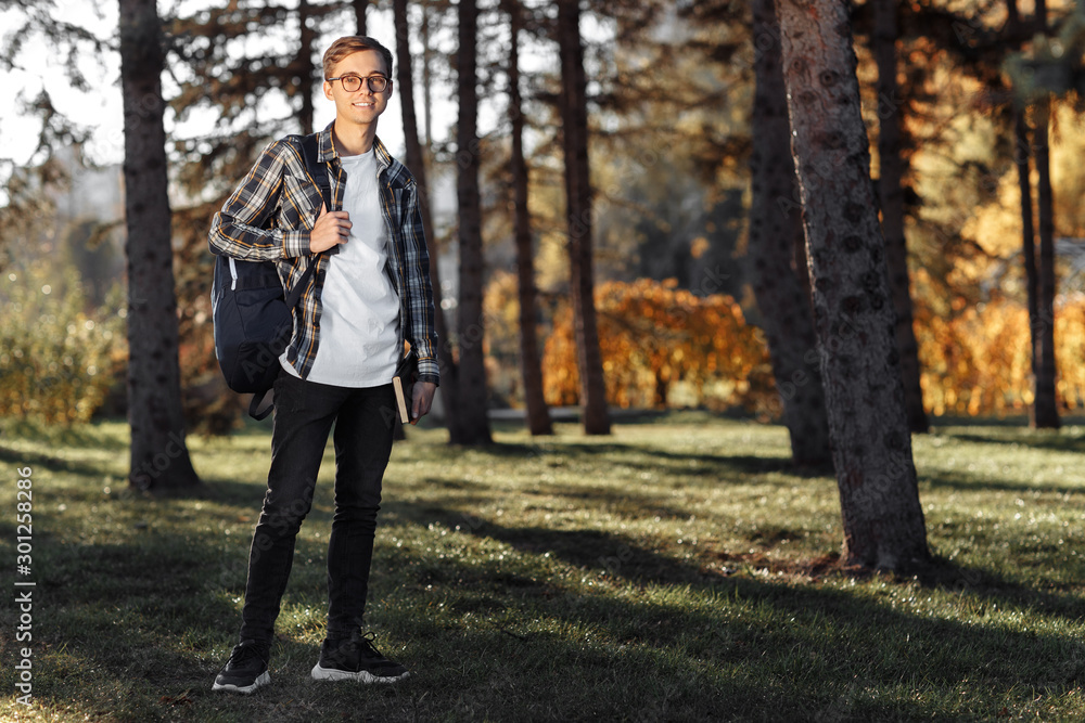 Full lengh photo of happy young male student with glasses in casual outfit holding backpack and posing at the park. Full lengh photo of happy young male student with glasses in casual outfit holding backpack and posing at the park.