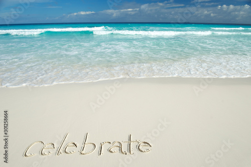 Celebrate message handwritten on a deserted tropical beach with blue waves crashing above smooth sand copy space