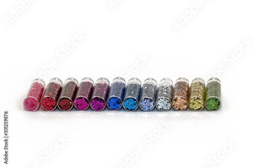 Colorful glitter particles for crafting or nail art design in transparent vials lying on white background