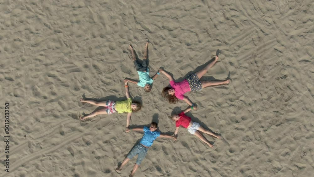 Five children in bright clothes lie on the sand in the shape of a star and wave their hands. Aerial shot of a friendly team of children Five children in bright clothes lie on the sand in the shape of a star and wave their hands. Aerial shot of a friendly team of children