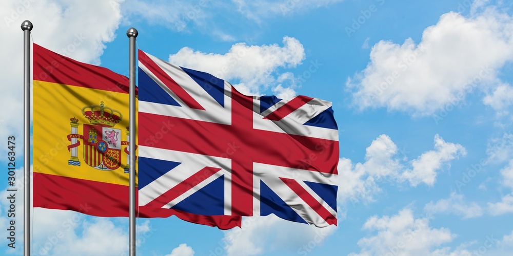 Spain and United Kingdom flag waving in the wind against white cloudy blue sky together. Diplomacy concept, international relations. Spain and United Kingdom flag waving in the wind against white cloudy blue sky together. Diplomacy concept, international relations.