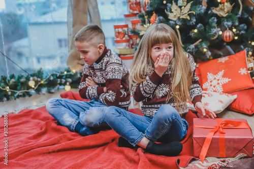 фотографія Greedy sister has stolen brother's present and looks at the camera with cunning face expression, while ffended boy is sitting near her looking upset