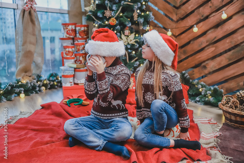 Full-body shot of beautiful children in santa hats and woolen sweaters, sitting near christmas tree. Boy is drinking a cocoa, his sister is looking at him with hungry face expression. photo