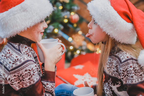 Tablou pe pânză Cropped shot of beautiful children in santa hats and woolen sweaters, sitting near christmas tree, with a cup of cocoa, looking delighted and pleased