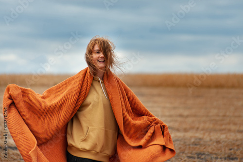 young girl with red hair and sun kissed face kidding on the yellow autumn field under dark cloudy sky wrapped in cosy orange plaid