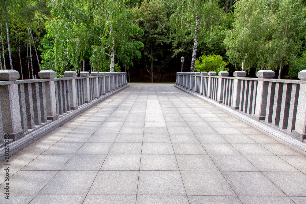 view of a bridge with stone tiles and granite railings with square columns in a park with trees, nobody.
