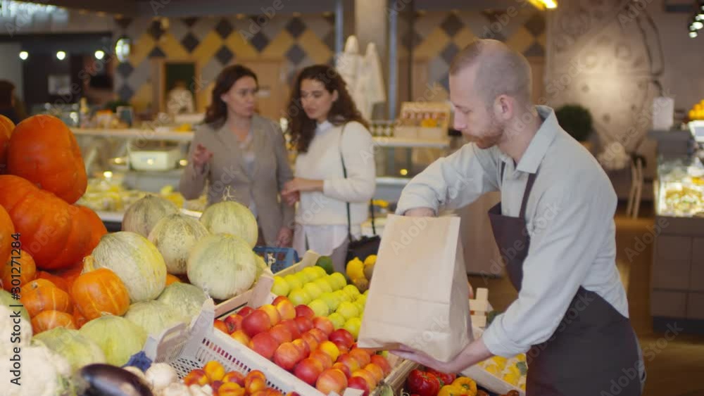 Salesman in apron putting fresh apples from craft paper bag into box on shelf while working in food market; two female customers choosing fruit in background Salesman in apron putting fresh apples from craft paper bag into box on shelf while working in food market; two female customers choosing fruit in background