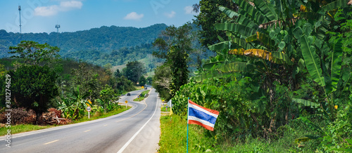 Asphalt road with Thai flags in a row, tropical forest and mountains near Khao Sok National Park, Thailand. It is the main tourist route to the Andaman sea - Phuket Island and Khao Lak.
