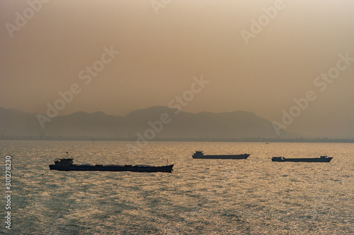 Da Nang, Vietnam - March 10, 2019: Tien Sa Port in Da Nang Bay. Three black coastal and river cargo vessels on orange light reflecting water of bay under sunset foggy sky 