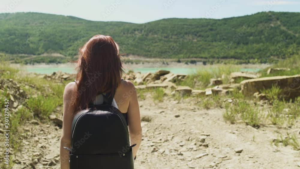 Beautiful confident redhead woman walking hiking at beautiful blue turquoise lake of Yesa, black backpack, with rocks and green forest background during sunny summer in Spain. Follow-Up 4K UHD. Beautiful confident redhead woman walking hiking at beautiful blue turquoise lake of Yesa, black backpack, with rocks and green forest background during sunny summer in Spain. Follow-Up 4K UHD.