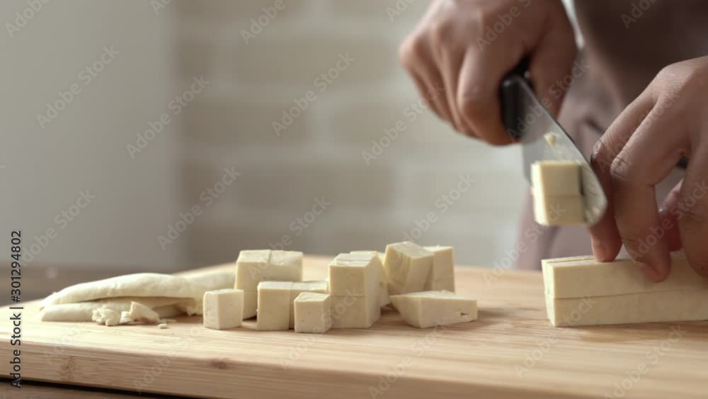 Closeup of home chef hands slicing and dicing tofu with kitchen knife on wooden cutting board preparing for cooking ingredient Closeup of home chef hands slicing and dicing tofu with kitchen knife on wooden cutting board preparing for cooking ingredient