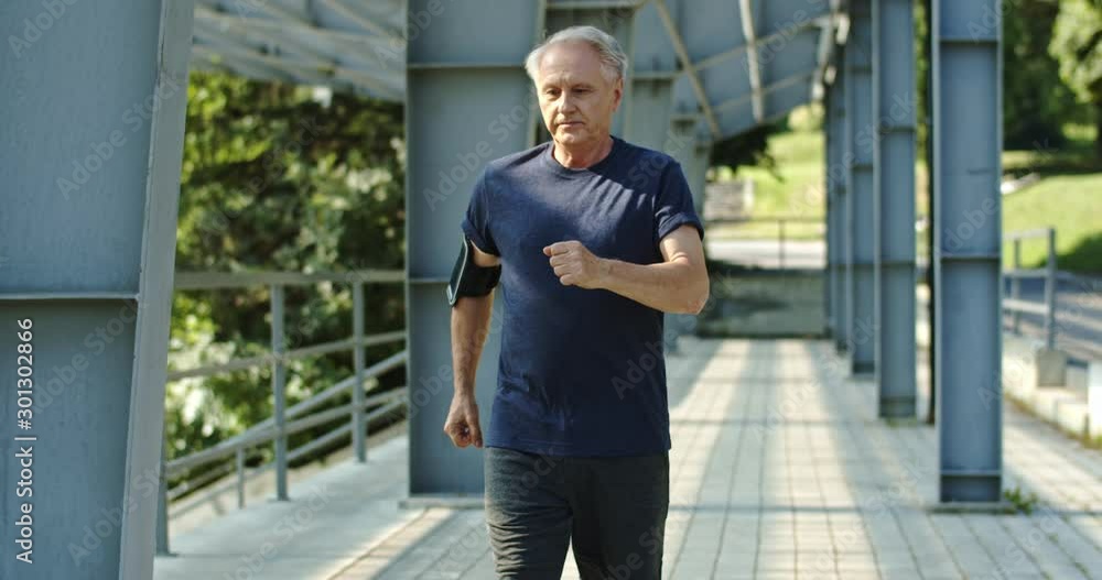 Strong senior Caucasian man in the good shape jogging in the morning at the stadium, then stops, breathing, having rest and keep on running.
