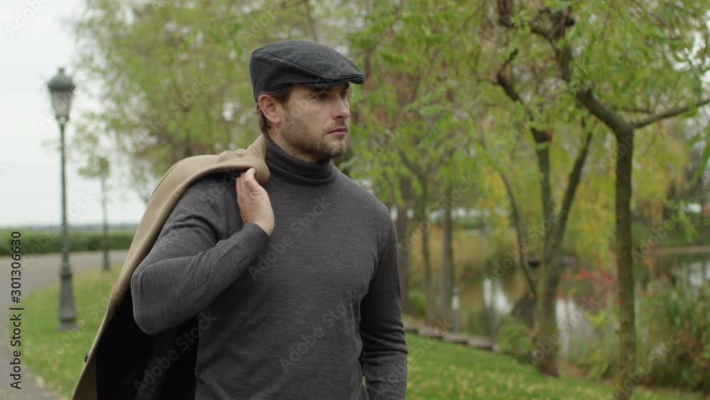 Good looking young European man walk in the park. Male takes off his beige coat and throws it on his shoulder. Good looking young European man walk in the park. Male takes off his beige coat and throws it on his shoulder.