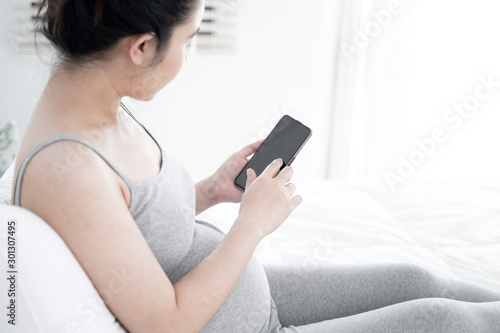 Young pregnant woman using smartphone with blank screen lying on bed. Young expectant lady surfing net, reading about baby care, buying goods for newborn concept.