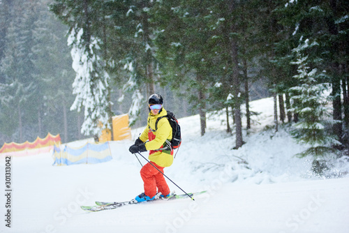 Male backpacker on skis in goggles and helmet in snowfalling weather, standing on steep wooded slope and posing on camera before skiing down at ski resort. Blurred background. Portrait horizontal shot