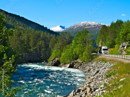 A truck is approaching on the road next to a rushing mountain stream that flows through rock and forest, with snow capped peaks and blue sky in the distance on a sunny summer day in Norway.