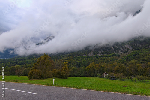 Scenic landscape photo of Slovenia. Small white house with front yard, valley with asphalt road, beautiful green meadow under mountain which shrouded by sick fog. Forest in the background
