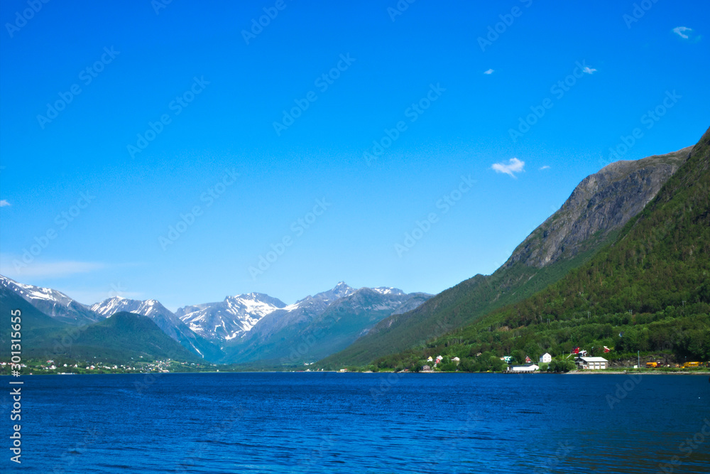 Isfjorden is the beautiful blue fjord at Andalsnes, Norway, surrounded by homes at the base of the mountains with snow and clear sky in the background on a sunny day.