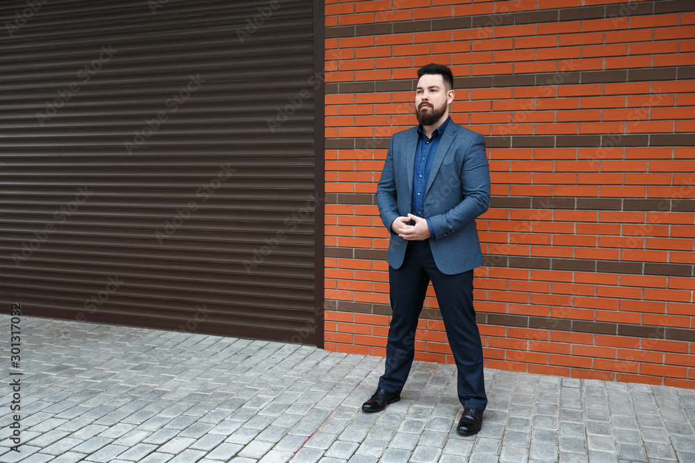 Stylish handsome brunette man with beard, wearing blue suit jacket, shirt, pants and shoes, standing outdoors on the city street near red brick wall. Classic elegant formal men's outfit. Businessman. Stylish handsome brunette man with beard, wearing blue suit jacket, shirt, pants and shoes, standing outdoors on the city street near red brick wall. Classic elegant formal men's outfit. Businessman.