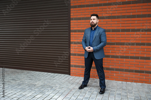 Stylish handsome brunette man with beard, wearing blue suit jacket, shirt, pants and shoes, standing outdoors on the city street near red brick wall. Classic elegant formal men's outfit. Businessman.
