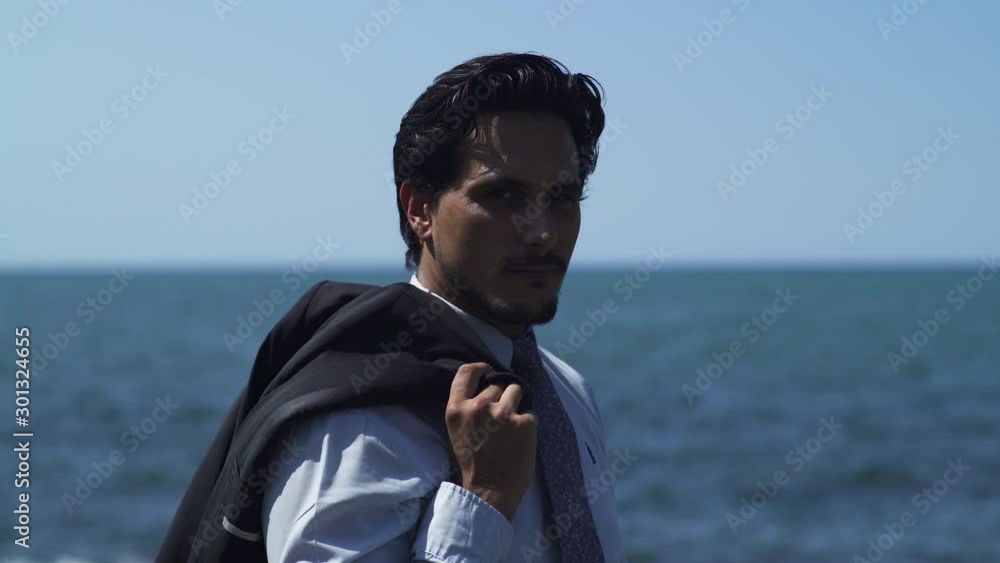 Businessman standing on a rock with blue sea at the beach in sunny summer, tie suit. Black sunglasses. Looking at camera thoughtful, determined and relaxed after work. Close-up 4K UHD. Smiling. Businessman standing on a rock with blue sea at the beach in sunny summer, tie suit. Black sunglasses. Looking at camera thoughtful, determined and relaxed after work. Close-up 4K UHD. Smiling.