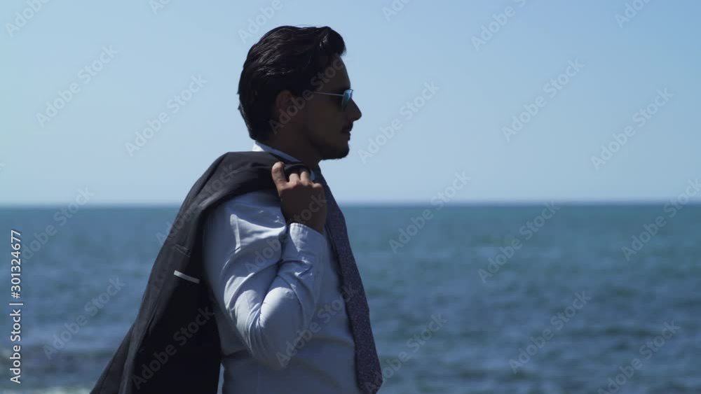 Businessman standing on a rock with blue sea at the beach in sunny summer, tie suit and leather shoes, black sunglasses. Looking away thoughtful, determined and relaxed after work. Medium shot 4K UHD. Businessman standing on a rock with blue sea at the beach in sunny summer, tie suit and leather shoes, black sunglasses. Looking away thoughtful, determined and relaxed after work. Medium shot 4K UHD.