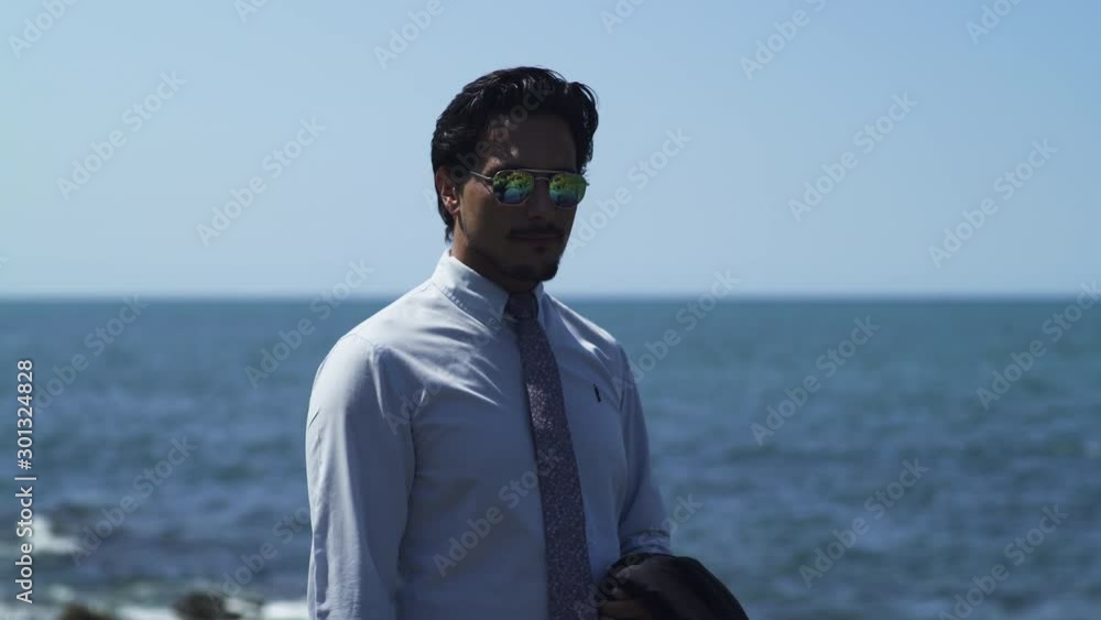 Businessman standing on a rock with blue sea at the beach in sunny summer, tie suit. Black sunglasses. Looking at camera thoughtful, determined and relaxed after work. Close-up 4K UHD. Steadicam move. Businessman standing on a rock with blue sea at the beach in sunny summer, tie suit. Black sunglasses. Looking at camera thoughtful, determined and relaxed after work. Close-up 4K UHD. Steadicam move.