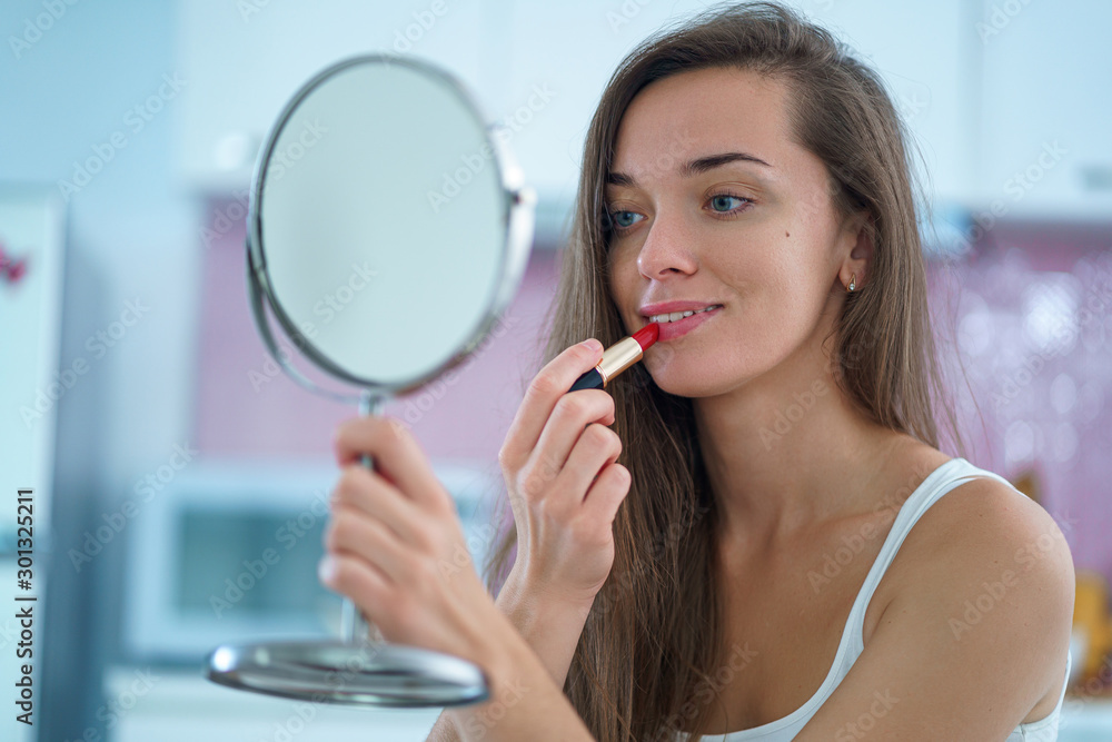 Beautiful attractive happy smiling brunette woman paints lips with red lipstick using a small round mirror during home makeup in the morning - obrazy, fototapety, plakaty 