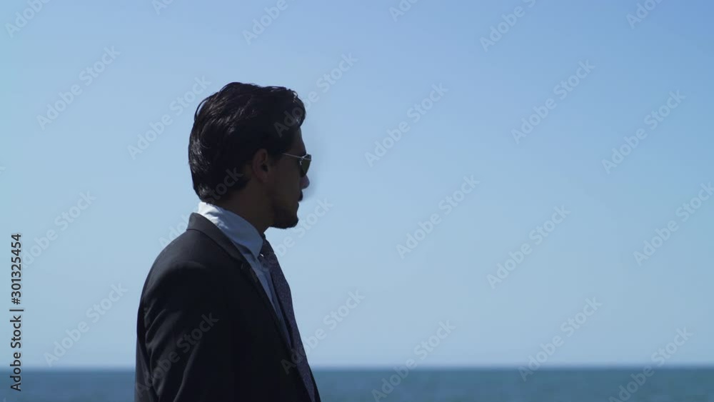 Businessman standing on a rock with blue sea at the beach in sunny summer, tie suit and leather shoes, black sunglasses. smoking cigarette after work. Wide Shot 4K UHD. Close-up. Businessman standing on a rock with blue sea at the beach in sunny summer, tie suit and leather shoes, black sunglasses. smoking cigarette after work. Wide Shot 4K UHD. Close-up.