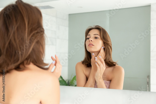 Beautiful girl with cream on face skin. Woman touching cheek with hand. Charming female looking at mirror. Home bathroom. Facial health. Skincare and hygiene. Daily makeup