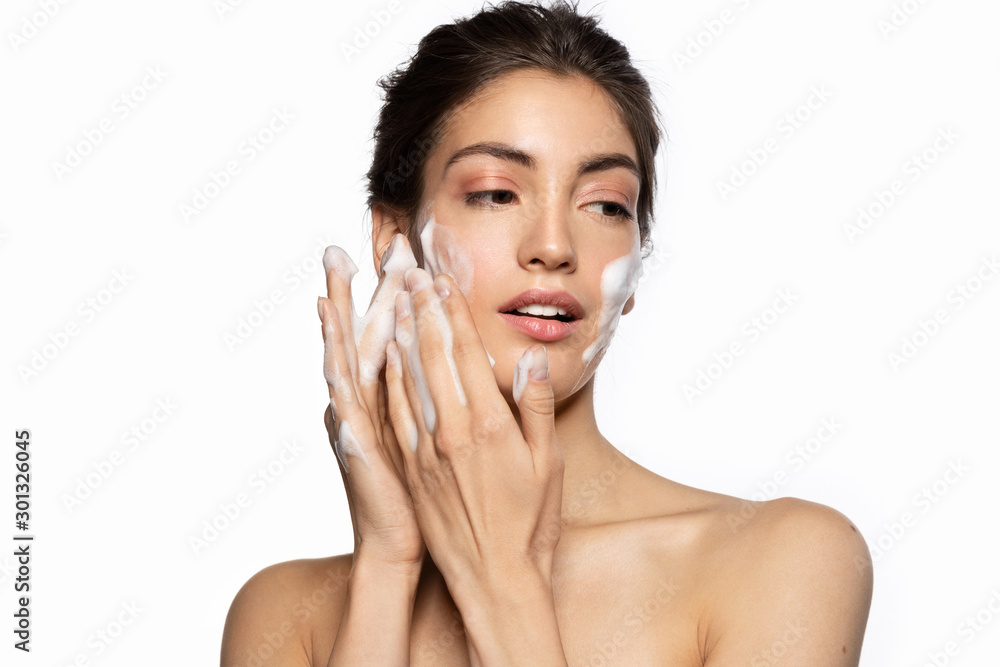 Beautiful young woman applying foam cleanser portrait. Pretty lovely brunette girl looking aside and touching cheek with soapy hands. Face cleansing and moisturizing. Studio white copy space Beautiful young woman applying foam cleanser portrait. Pretty lovely brunette girl looking aside and touching cheek with soapy hands. Face cleansing and moisturizing. Studio white copy space