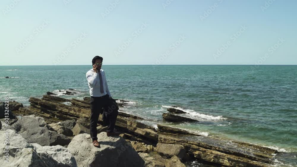 Businessman standing on a rock with blue sea at the beach in sunny summer, tie suit and leather shoes, black sunglasses. Having phone conversation. Wide Shot 4K UHD. Businessman standing on a rock with blue sea at the beach in sunny summer, tie suit and leather shoes, black sunglasses. Having phone conversation. Wide Shot 4K UHD.