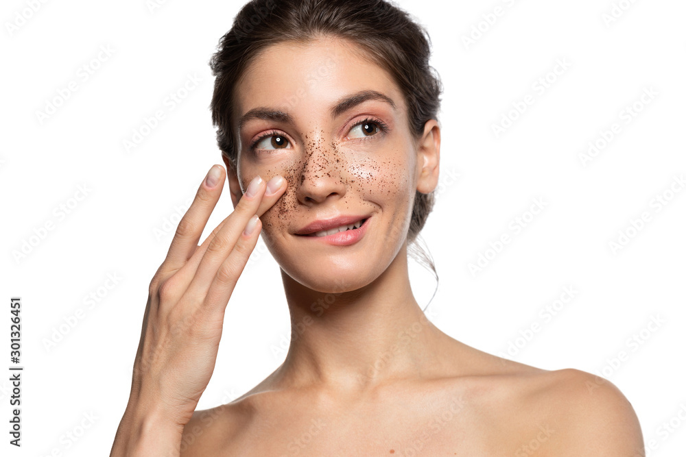 Close up portrait of slyly smiling woman applying cosmetic facial skin scrub, natural coffee mask. Beautiful young happy brunette girl cleaning face with beauty product. White studio blank copy space