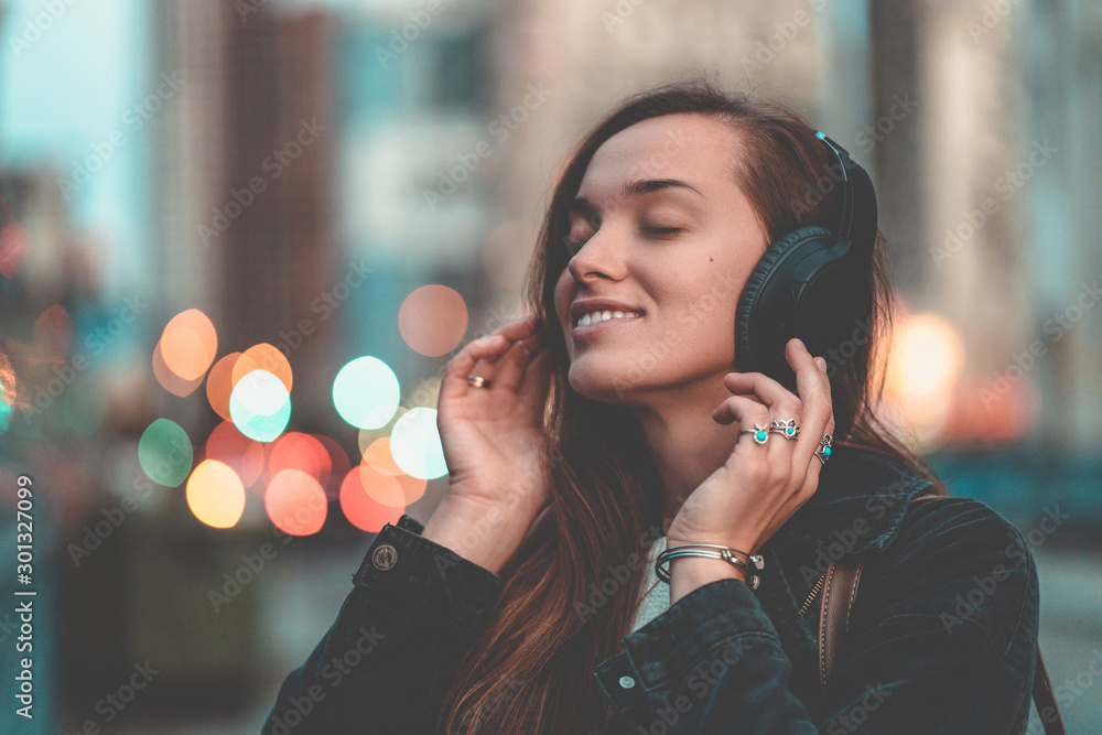 Young happy stylish trendy casual hipster woman teenager listening to music on a wireless headphone while walking around the city. Music lover enjoying music - obrazy, fototapety, plakaty 