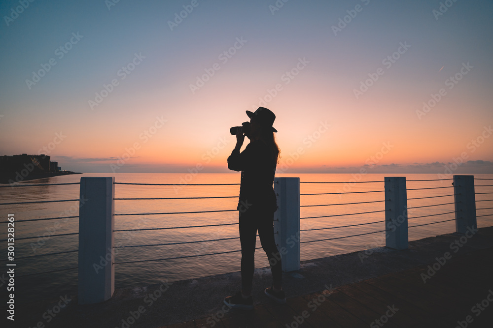 Silhouette of woman photographer in hat with digital camera during taking photos of sea at pier at sunset