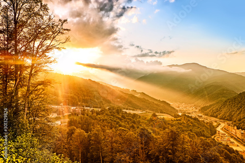 sunset on mountains valley landscape against beautiful clouds sky background. Aerial wide view of mountain nature forest and town with river in orange setting sun light. Idyllic evening wallpaper